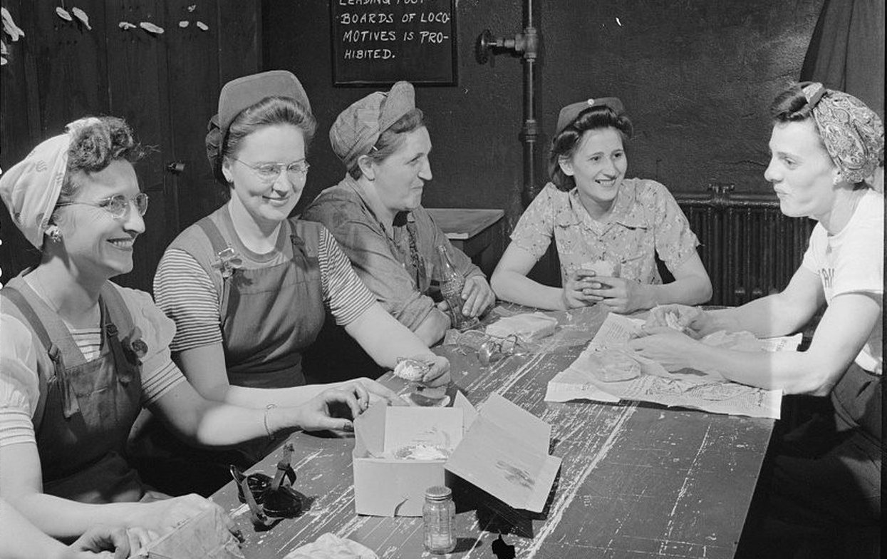 Five women railroad workers laughing over lunch at a long wooden table